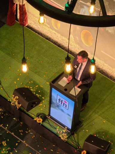 Mann am Rednerpult auf einer Bühne mit dekorativen Lichtern und farbigen Blüten.